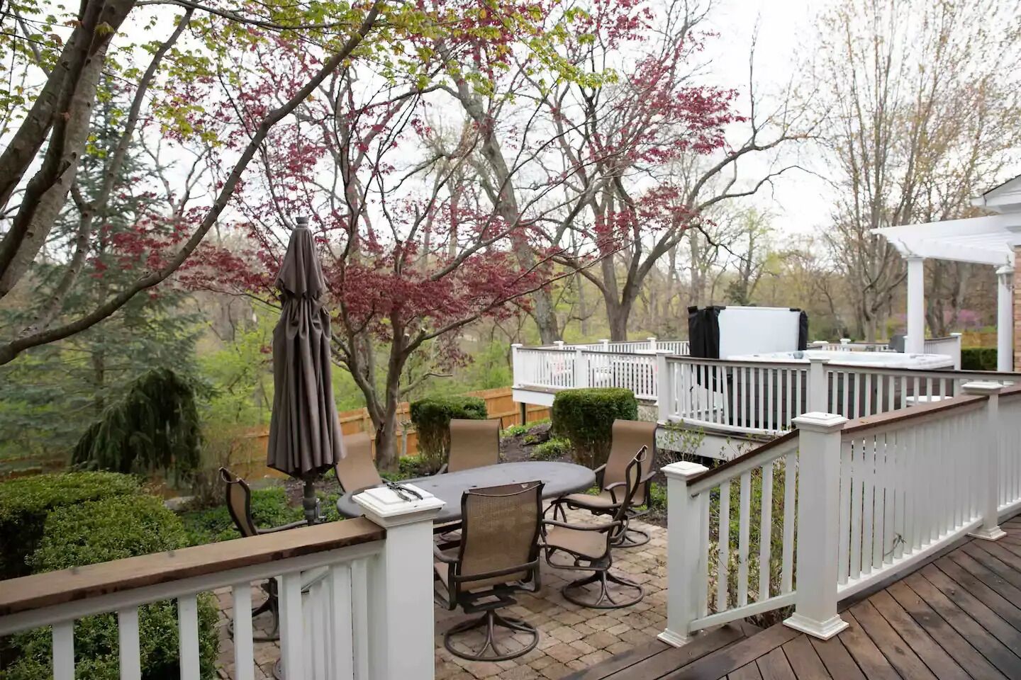 A covered outdoor patio with a table and chairs, surrounded by a white fence and trees with pink leaves.