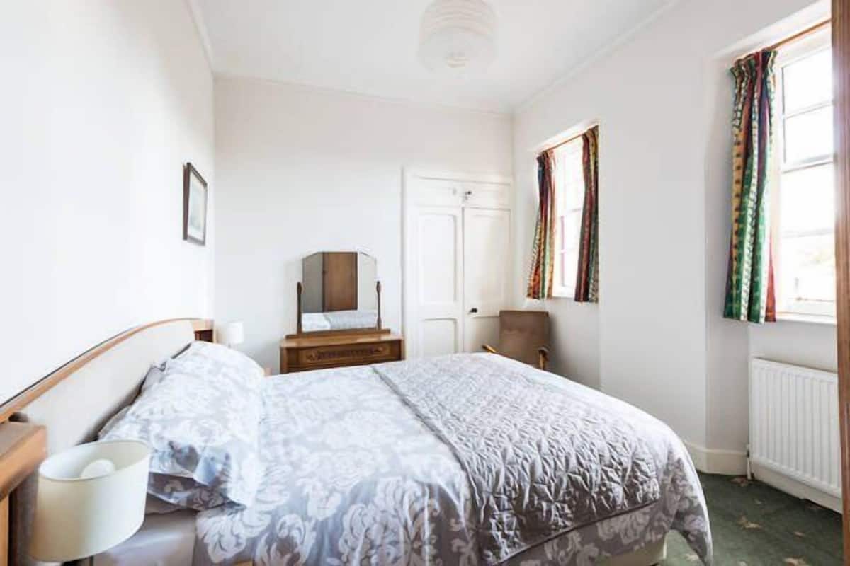 A bedroom with a bed, a bedside table, a mirror, a dresser, and a window with curtains.