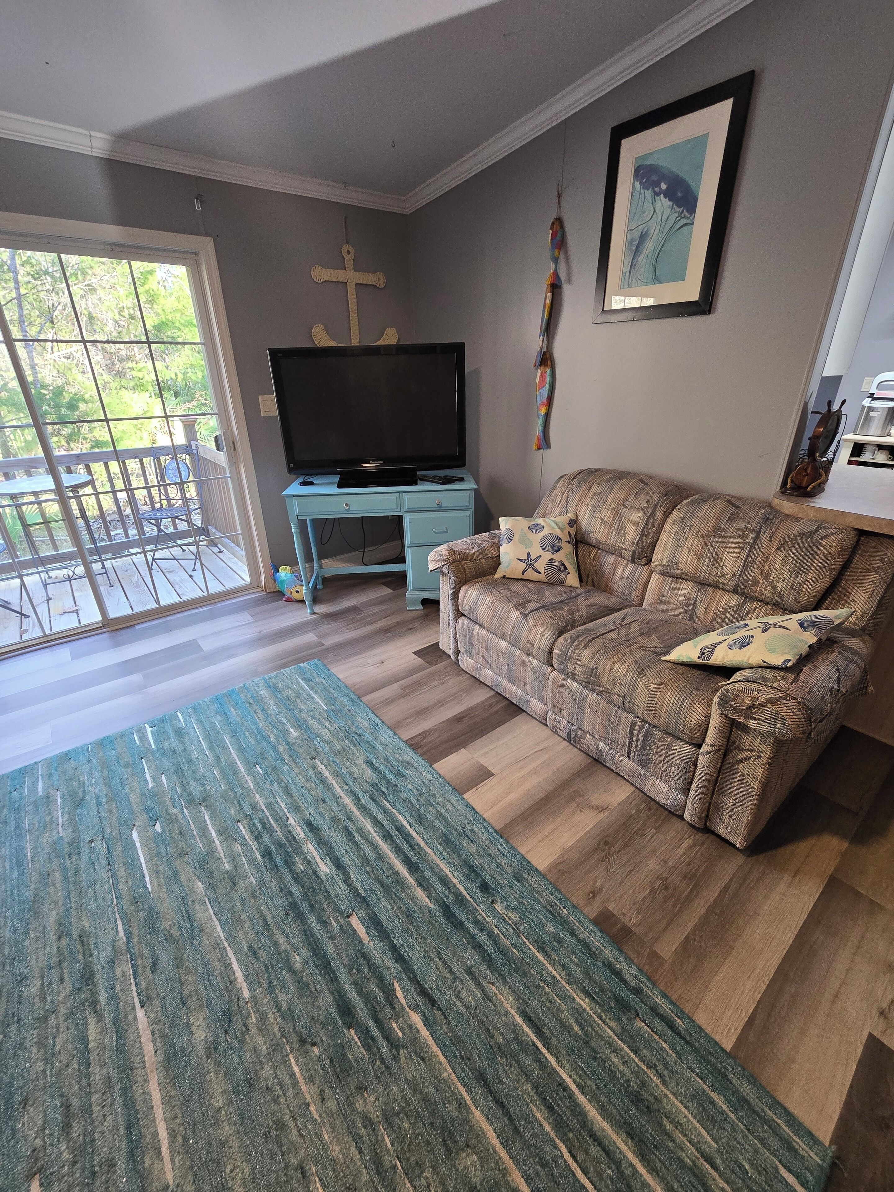 A living room with a sofa, a television on a stand, a patterned rug, and a view through a glass door to an outdoor area with a railing.