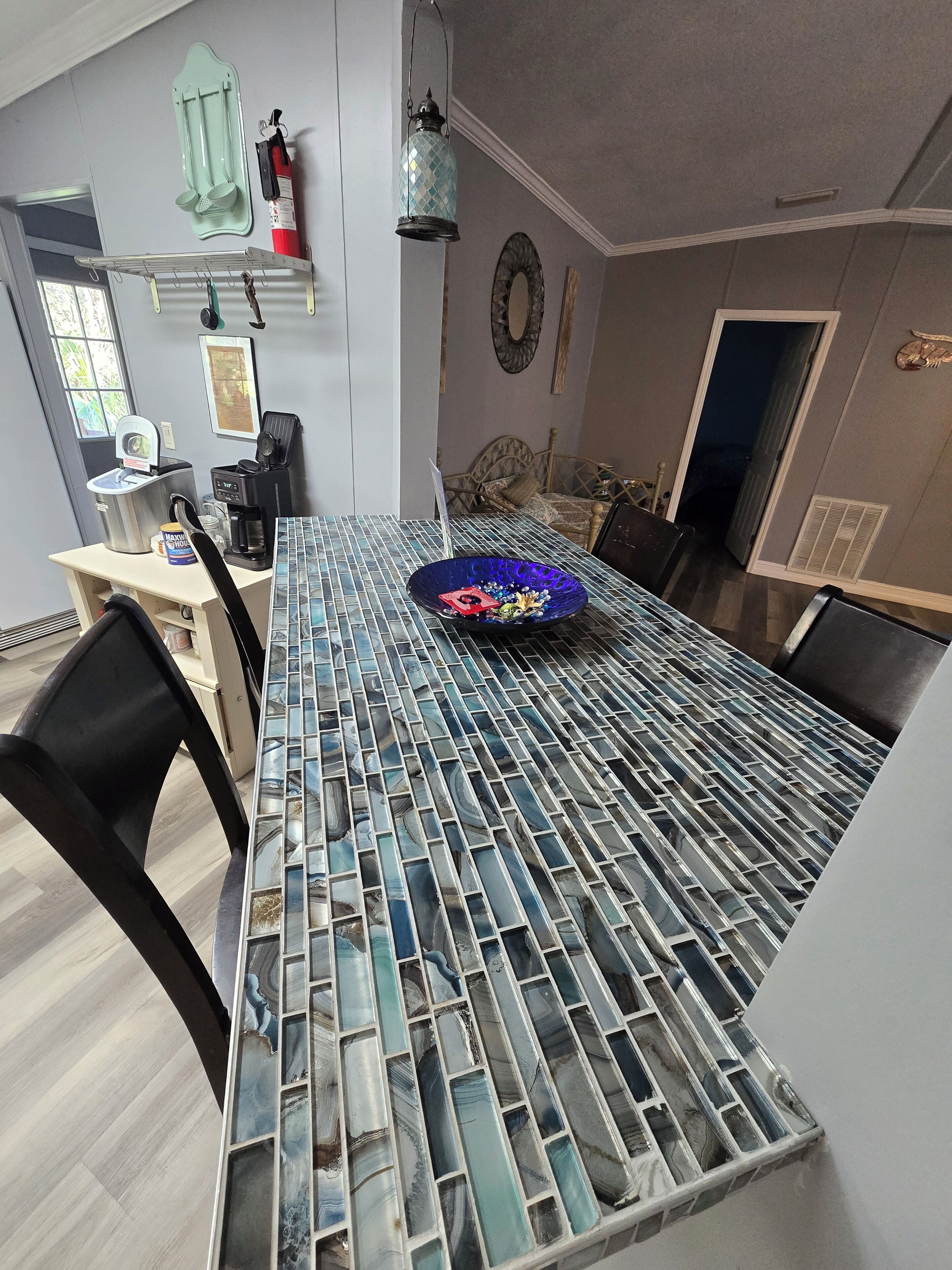 Un espace repas comprenant une table avec un plateau en verre, des chaises, une machine à café, un extincteur et un miroir décoratif.
