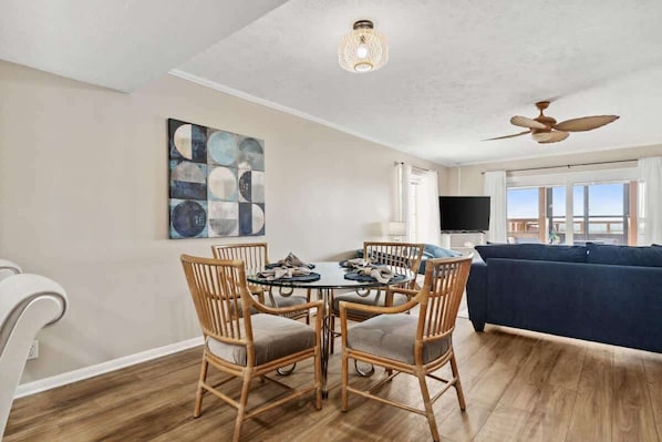 Un coin repas avec une table ronde et des chaises, un ventilateur de plafond, un téléviseur et un canapé, dans une pièce dotée d’un plancher en bois et d’une grande fenêtre.