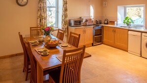 A dining area with a wooden table set for four, a clock on the wall, and a kitchen with wooden cabinets and appliances. - Wynford Holiday Cottages (Aberdeen)