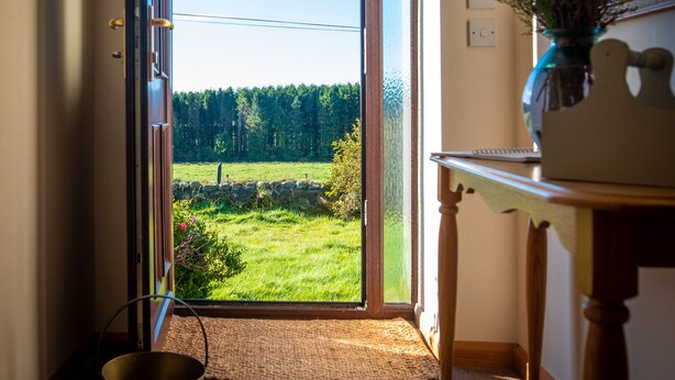 A room with a large glass door leading to a grassy outdoor area, a small table with a vase, and a metal bucket on the floor. - Wynford Holiday Cottages (Aberdeen)