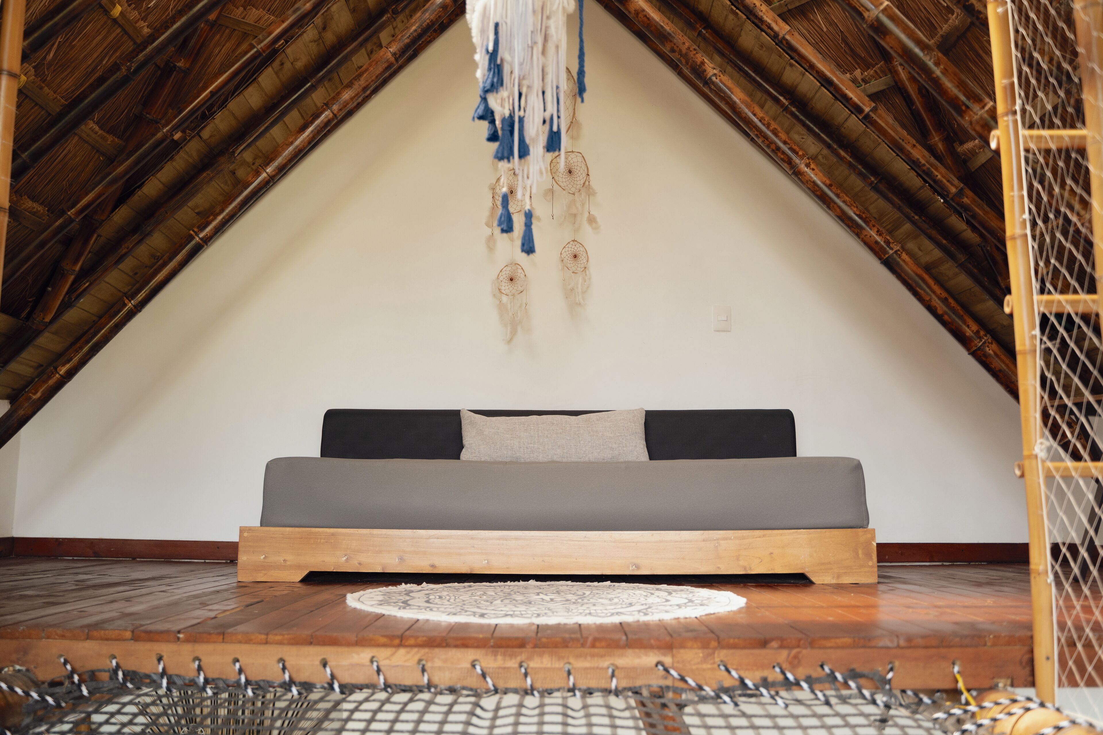 A loft bedroom with a wooden ceiling, a gray sofa, and a hanging dreamcatcher.