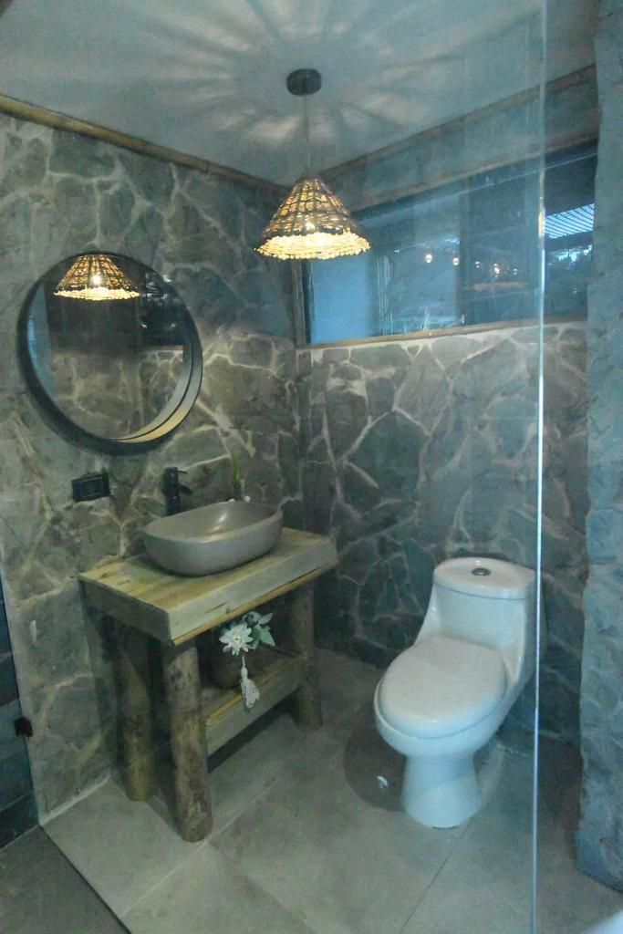 Un baño con pared de piedra, tocador de madera, espejo redondo, lavabo sobre encimera, inodoro y lámpara colgante.
