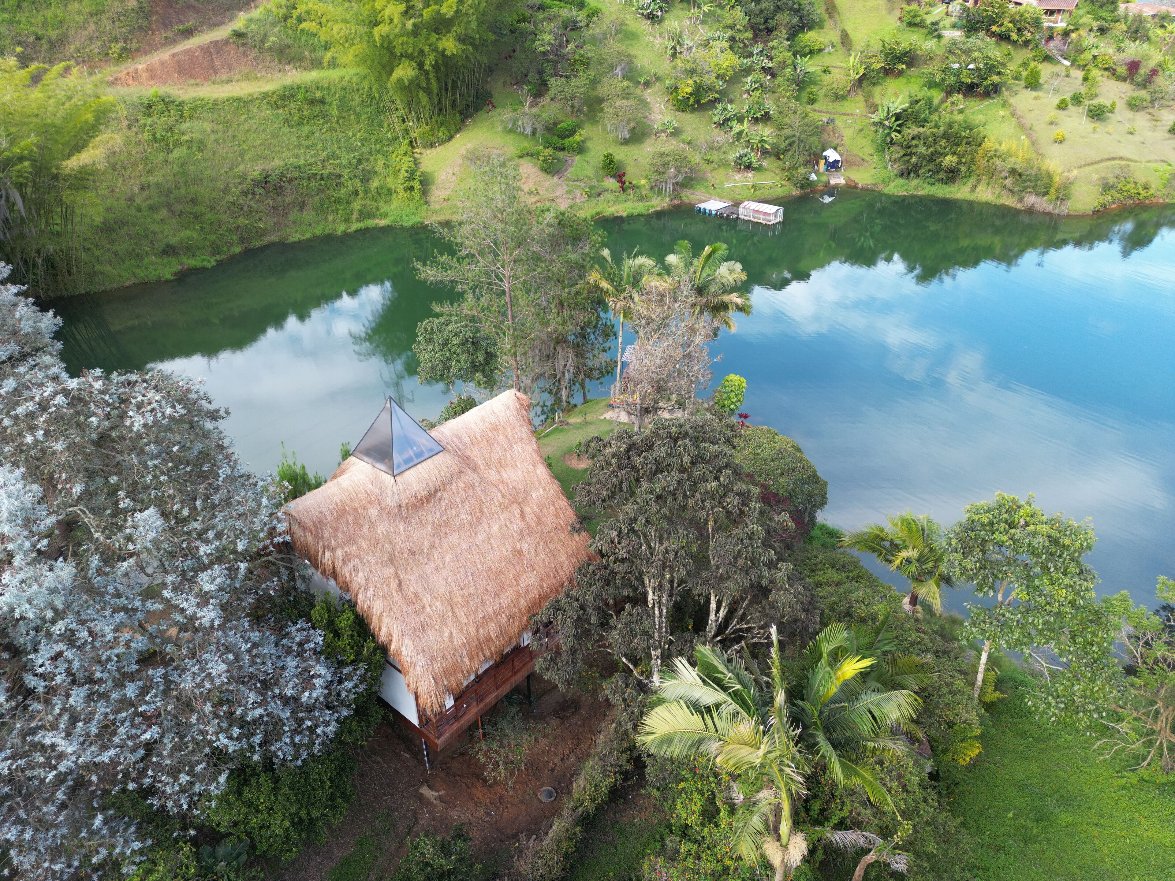 A thatched-roof hut with a small attached structure, surrounded by lush greenery and a calm lake.