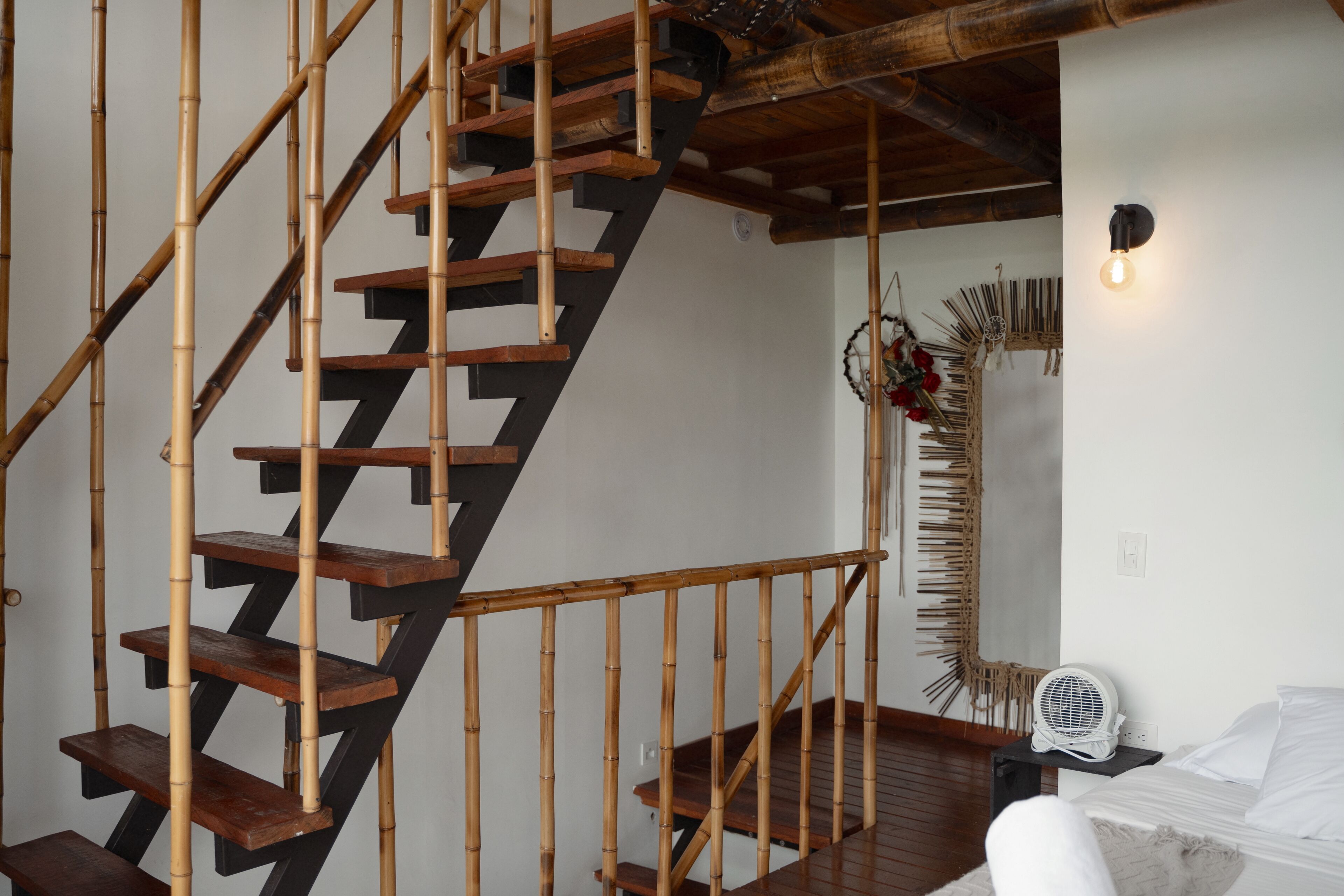 Una escalera con escalones de madera y barandilla de bambú, una cama con ropa blanca, un ventilador fijado a la pared y un adorno mural.