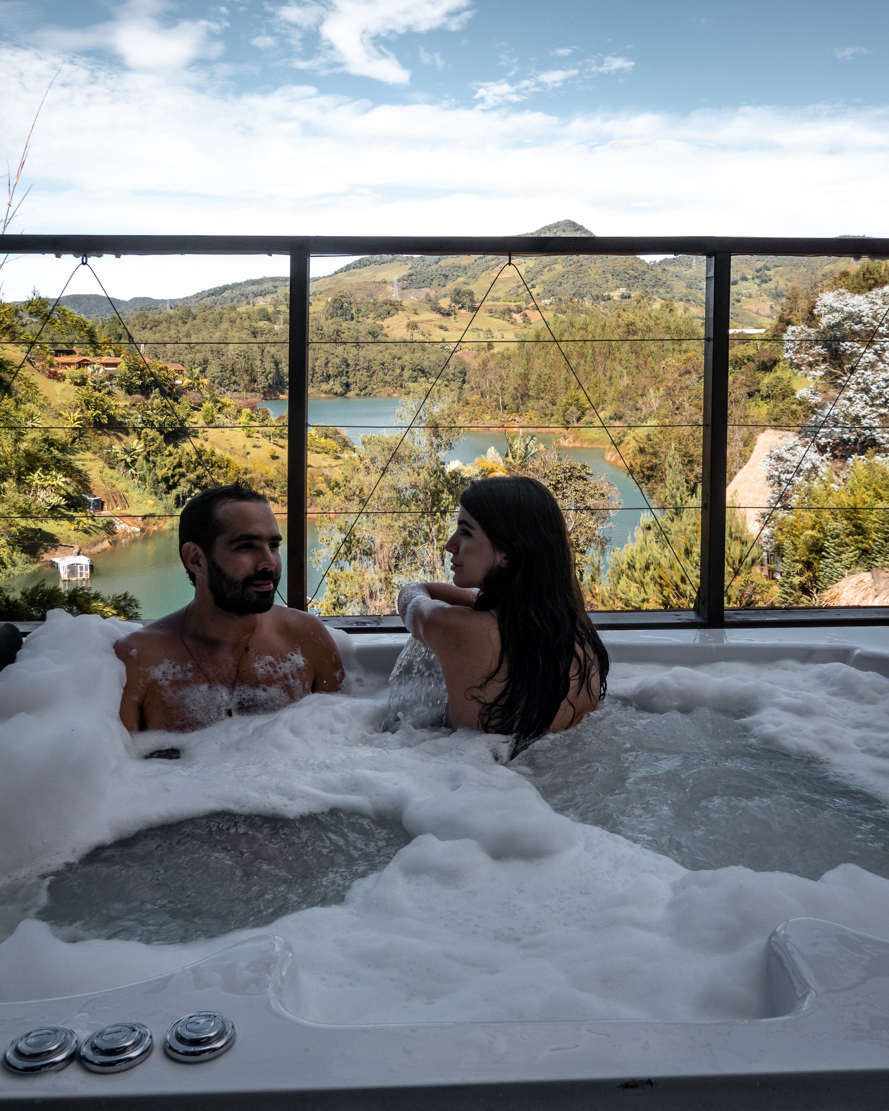 A couple in a hot tub with a scenic view of hills and a lake.