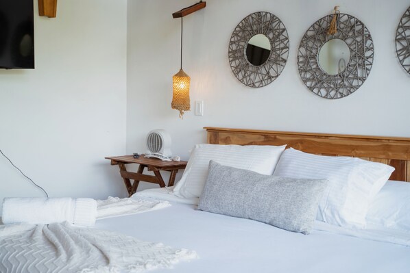 A neatly made bed with white linens and a grey pillow, a wooden bedside table with a fan, and two wall-mounted mirrors with a woven light fixture above.