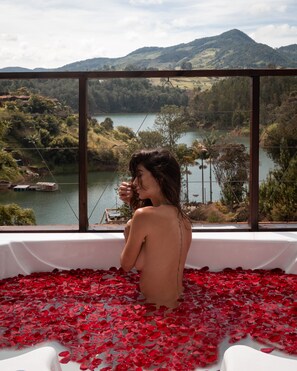 A bathtub filled with rose petals, a wooden railing, and a view of a lake and hills.