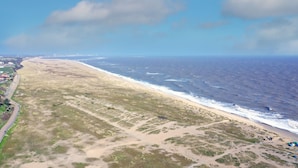 Eine Vogelperspektive zeigt einen Strand mit einer parallel zur Küste verlaufenden Straße, einen klaren Himmel und das Meer.