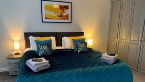 Cama bem arrumada com edredom verde-água, duas almofadas e uma toalha dobrada em cada lado. Há uma obra de arte emoldurada acima da cama e um guarda-roupa à direita.