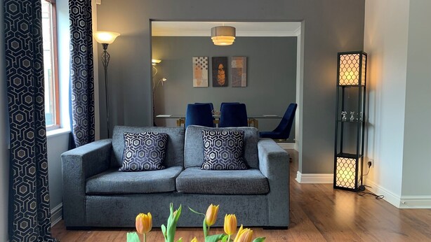 Uma sala de estar com um sofá cinza, almofadas estampadas, um tapete azul e um vaso de tulipas.