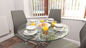 Uma mesa de jantar com quatro cadeiras, tampo de vidro e uma mesa posta para o café da manhã, incluindo pratos, tigelas, copos e suco de laranja.