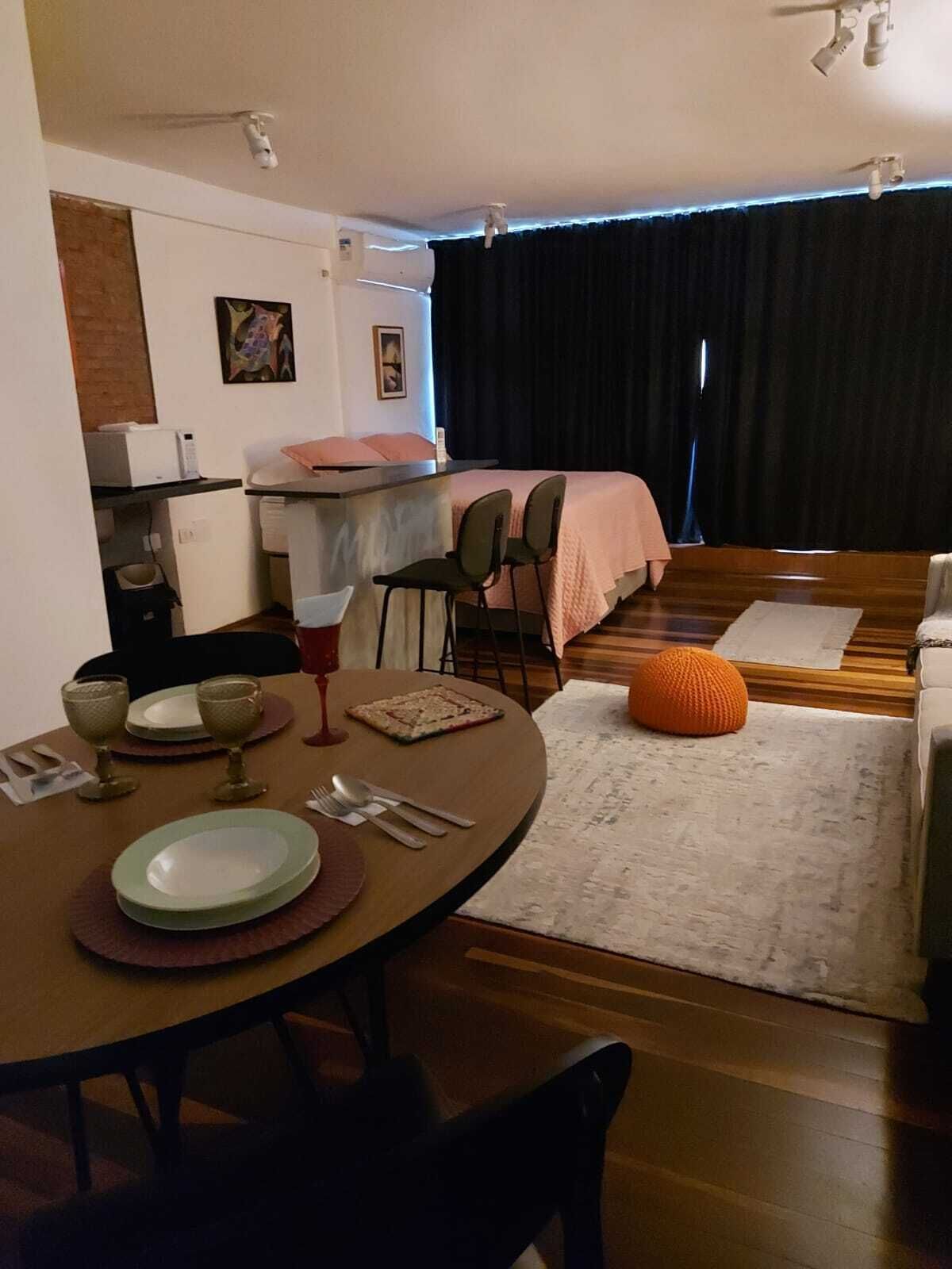 Un espacio habitable compacto con comedor, kitchenette y cama con cortina oscura de fondo.
