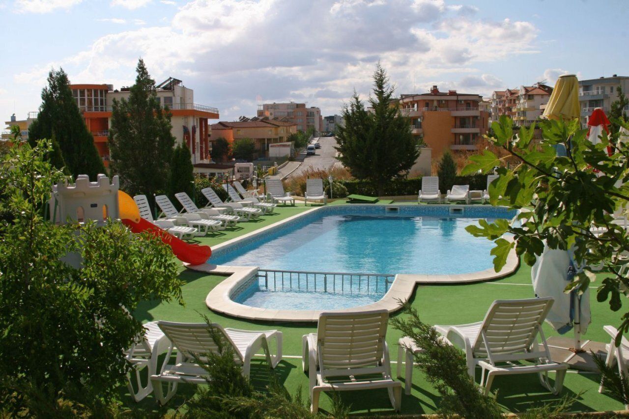 Une piscine dotée de chaises longues et d’un toboggan, entourée de verdure et de bâtiments.