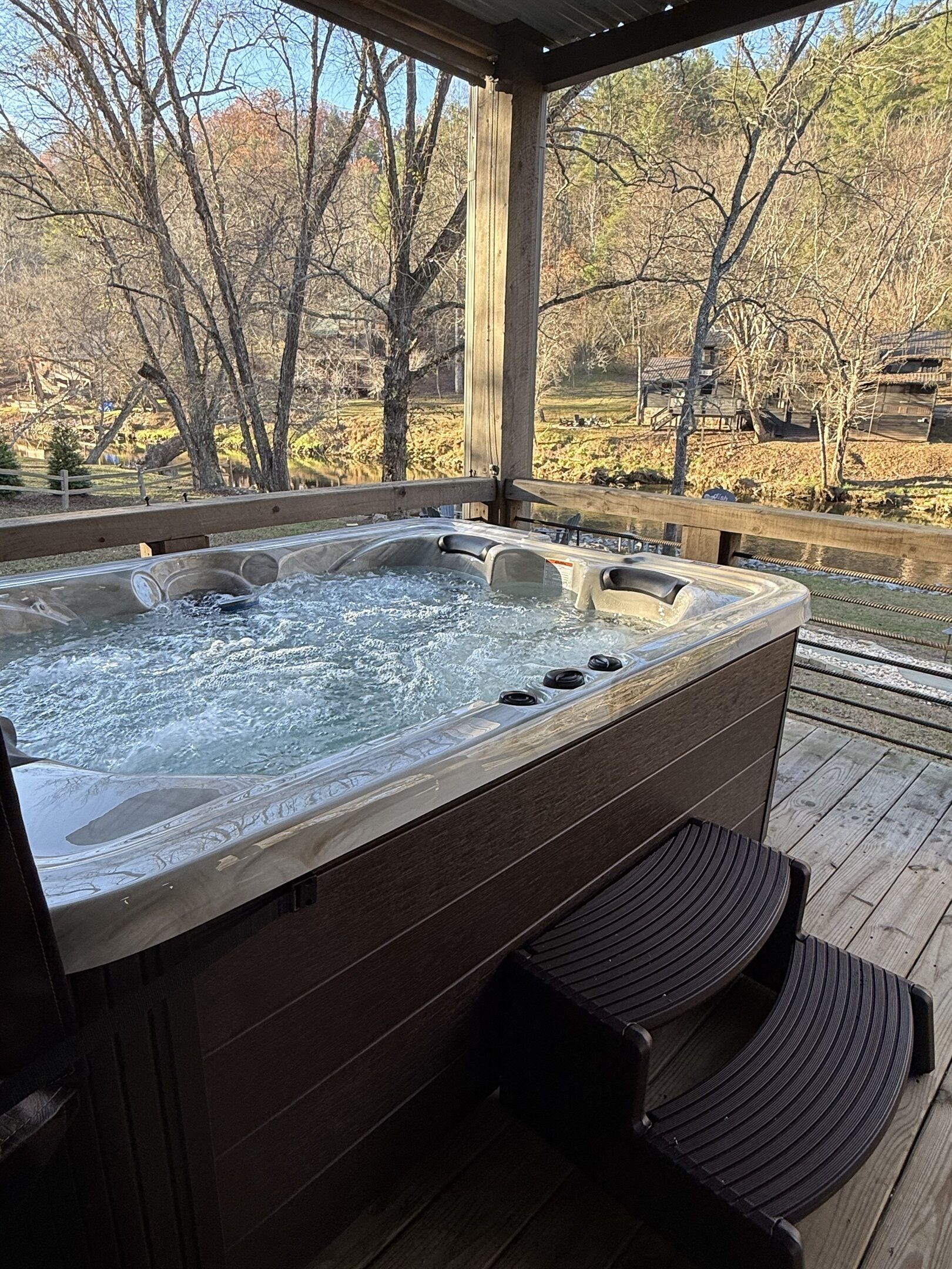 Ein Whirlpool mit einer Holzterrasse und Stufen, umgeben von Bäumen und einem Holzzaun.