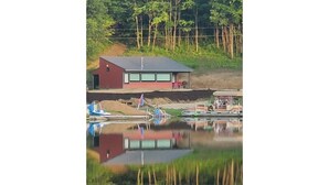 A house with a red exterior and a grey roof, situated next to a calm body of water reflecting the structure. - Move-in ready lakeside retreat (Minerva)