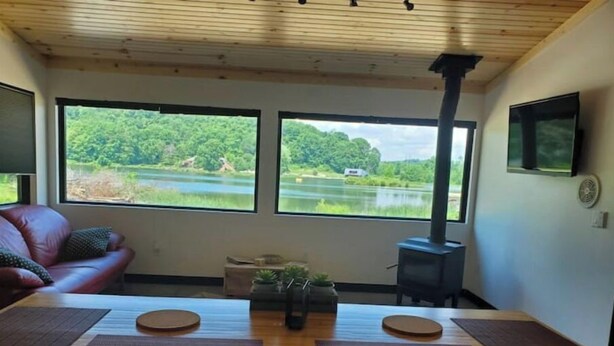 A cozy room with a wooden table, two windows offering a view of a lake, a red leather couch, and a wood-burning stove. - Move-in ready lakeside retreat (Minerva)