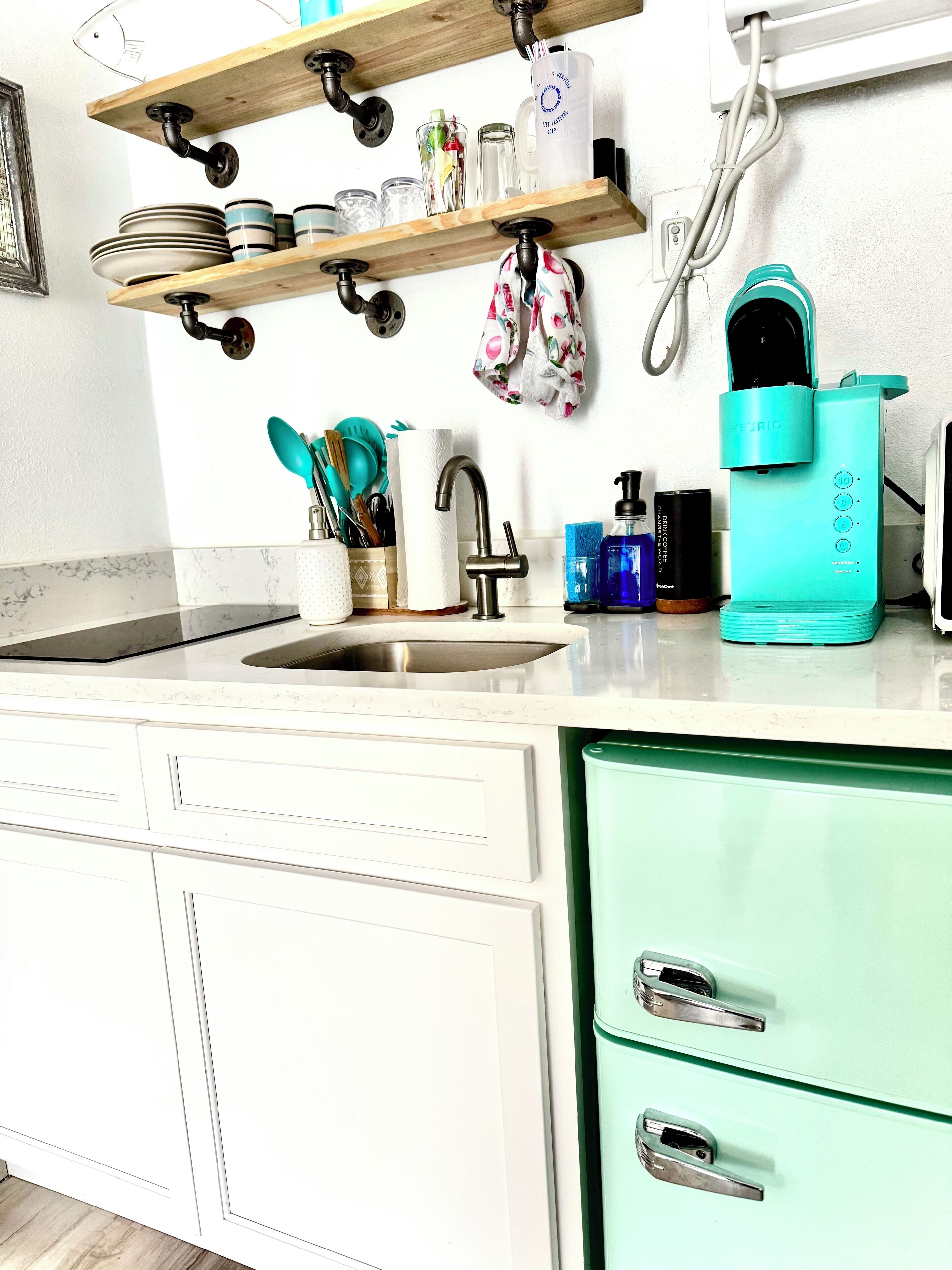 Une cuisine avec des meubles blancs, un évier, une cafetière turquoise et divers ustensiles de cuisine rangés sur des étagères.