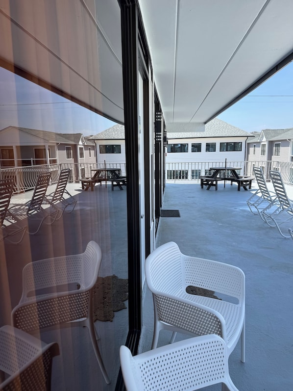 A covered outdoor seating area with white plastic chairs and tables, overlooking a row of white buildings.