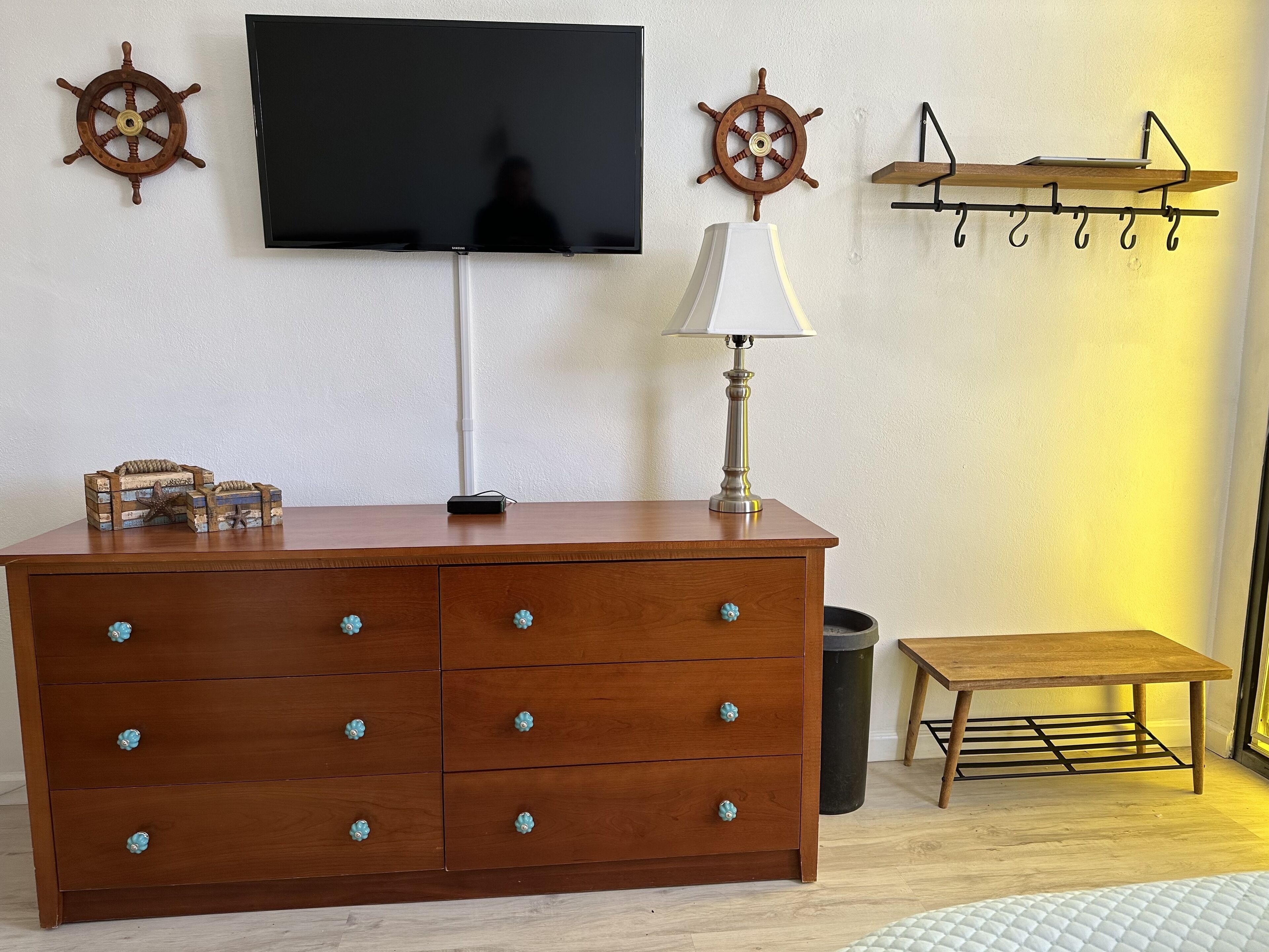 Une commode en bois avec des tiroirs, un téléviseur fixé au mur, une lampe et une petite table avec un présentoir en métal.