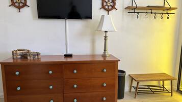 Une commode en bois avec des tiroirs, un téléviseur fixé au mur, une lampe et une petite table avec un présentoir en métal.
