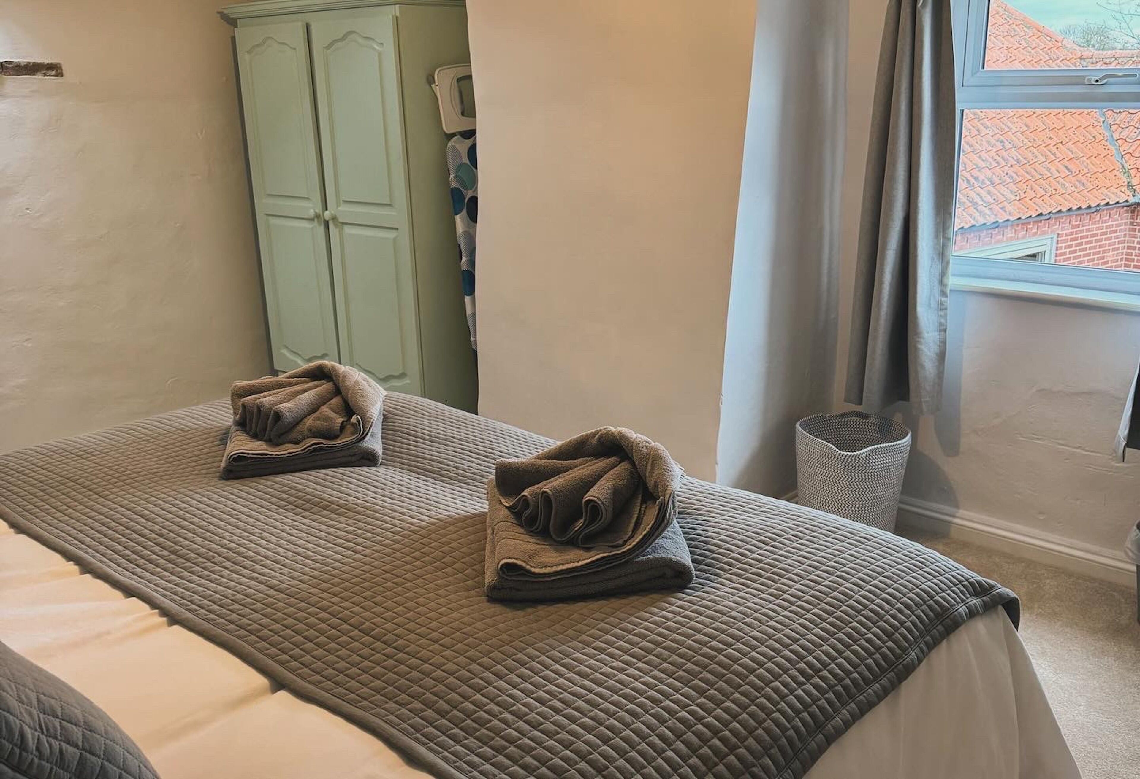 A hotel room with a bed, two towels on the bed, a wardrobe, a window with a view of a building, and a trash bin.