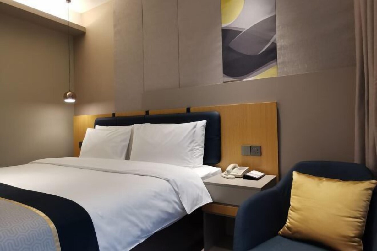 Ein Hotelzimmer mit einem ordentlich bezogenen Bett, einem Nachttisch mit Telefon und Lampe sowie einem Sessel mit Kissen.