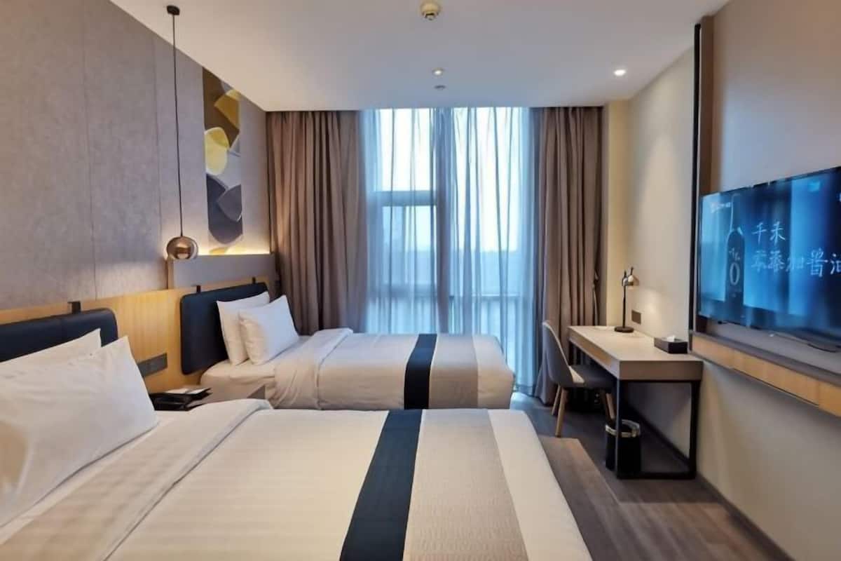 Habitación de hotel con dos camas, un escritorio, un televisor y un ventanal grande con cortinas.