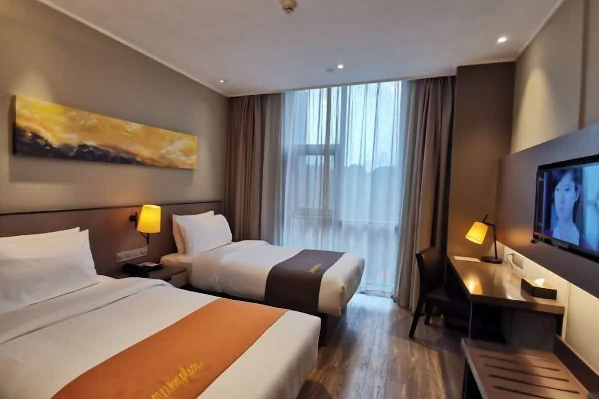 Habitación de hotel con dos camas, un escritorio con silla, un televisor y un ventanal con cortinas.