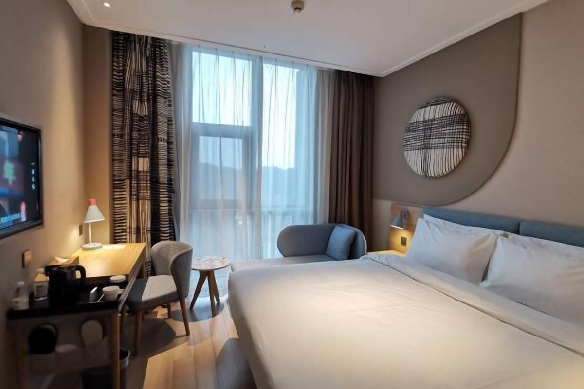 Una habitación de hotel con cama, escritorio, silla, televisor y un ventanal con cortinas.