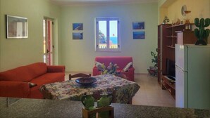 A compact living space with a dining area, a red sofa, a table with a floral tablecloth, and a refrigerator. - Villa Selenia, completamente autonoma a pochi metri dal mare! (JOPPOLO)