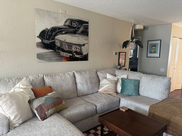 A living room with a grey sectional sofa, a wooden coffee table, and a large vintage car poster on the wall.