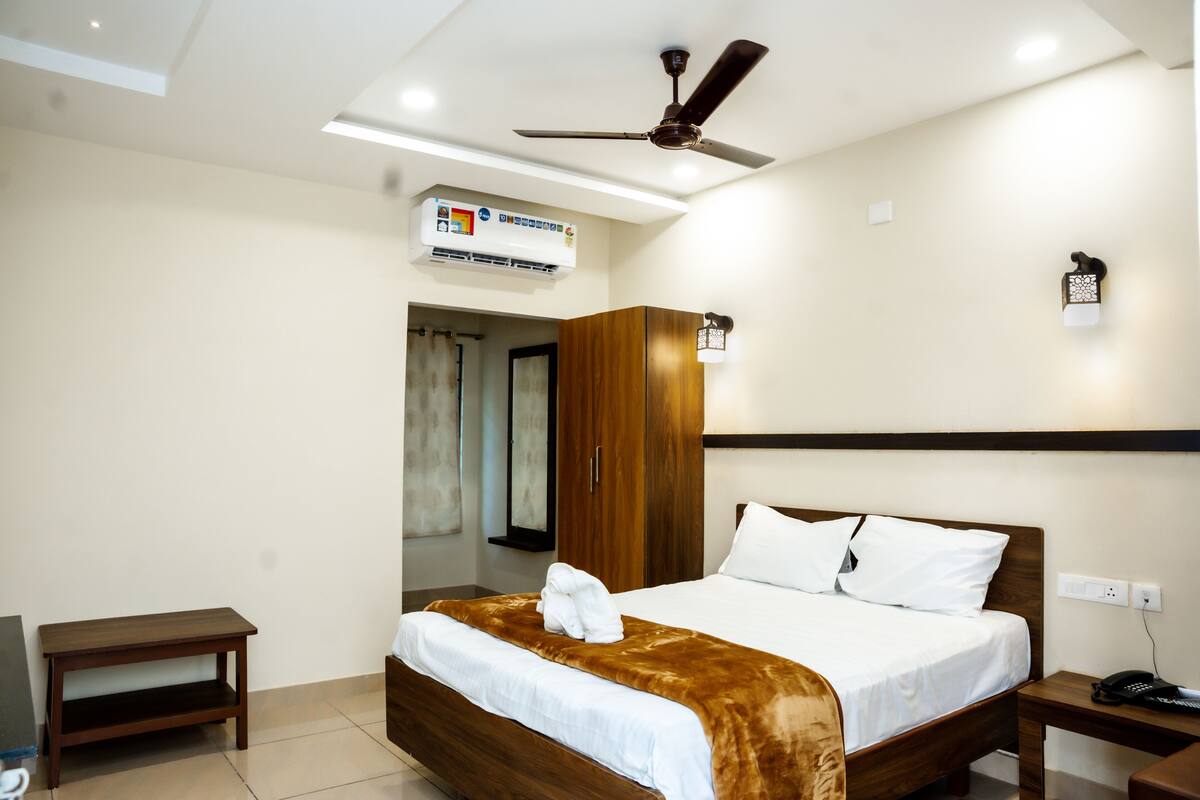 A hotel room with a bed, a bedside table, a ceiling fan, an air conditioning unit, and a nightstand with a phone.