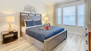 A modern bedroom with a wooden bed, two bedside tables with lamps, a decorative tree on the wall, and large windows with blinds.