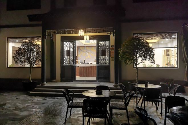 An outdoor seating area with tables and chairs, a potted tree, and a building entrance with decorative elements.