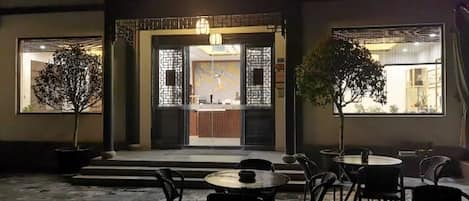 An outdoor seating area with tables and chairs, a potted tree, and a building entrance with decorative elements.