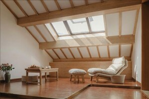 Une chambre à coucher aménagée dans un grenier, avec un plafond en pente, un puits de lumière, un fauteuil blanc rembourré, une petite table et une plante en pot.