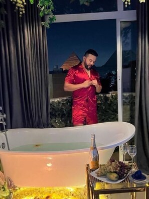 A man in a red bathrobe standing by a large bathtub with a tray of fruits and a bottle of champagne. - Pyramids view apartment with hot tub jacuzzi (Giza)