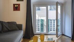 Een woonkamer met een bank, een tafel met twee mokken en een boek, een schuifdeur naar een balkon met groene luiken, en twee decoratieve wandpanelen.