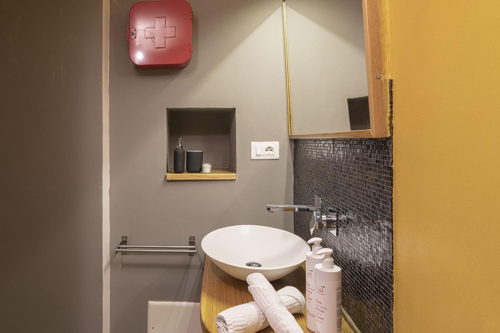 A bathroom with a white sink, a mirror, a red first aid kit, and a shelf with toiletries.