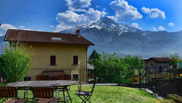 Une maison jaune avec un toit en tuiles, une terrasse avec des meubles en bois, et une montagne en arrière-plan.
