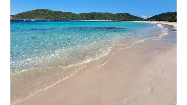 Une plage à l’eau turquoise et limpide, bordée d’un rivage sablonneux, avec des collines verdoyantes en arrière-plan.