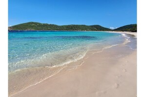 Une plage à l’eau turquoise et limpide, bordée d’un rivage sablonneux, avec des collines verdoyantes en arrière-plan.