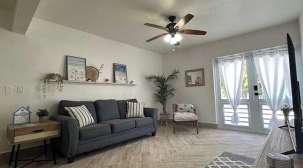 Un salon avec un canapé gris, une table d’appoint en bois, un ventilateur de plafond et une grande fenêtre avec des rideaux.