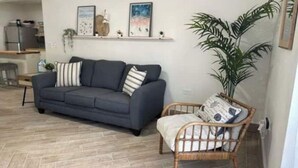 Un salon avec un canapé gris, un fauteuil en osier, une grande plante et, au fond, une cuisine.
