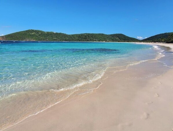 Een strand met helder turquoise water, een zandstrand en groene heuvels op de achtergrond.