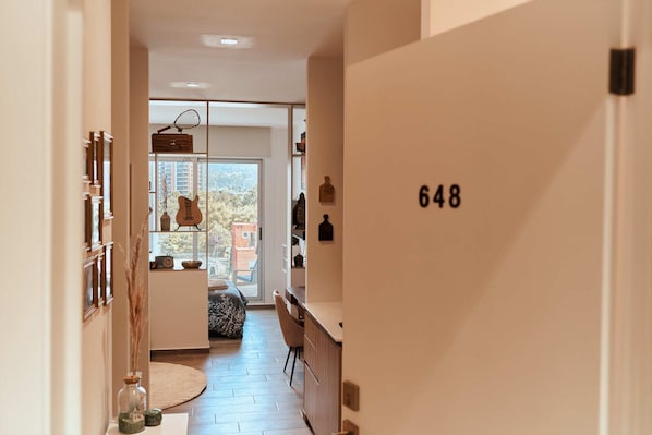 A modern interior with a wooden door marked 648, a kitchen area with a dining table, chairs, and a view through a glass door to an outdoor balcony.
