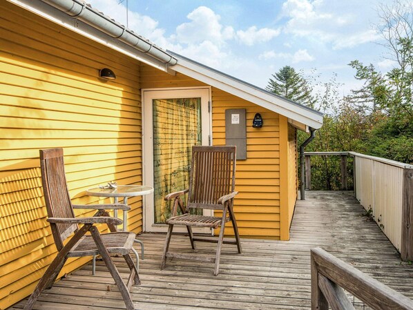 A wooden deck with two chairs and a small table, a door with a window, and a mailbox on the yellow siding of a building.